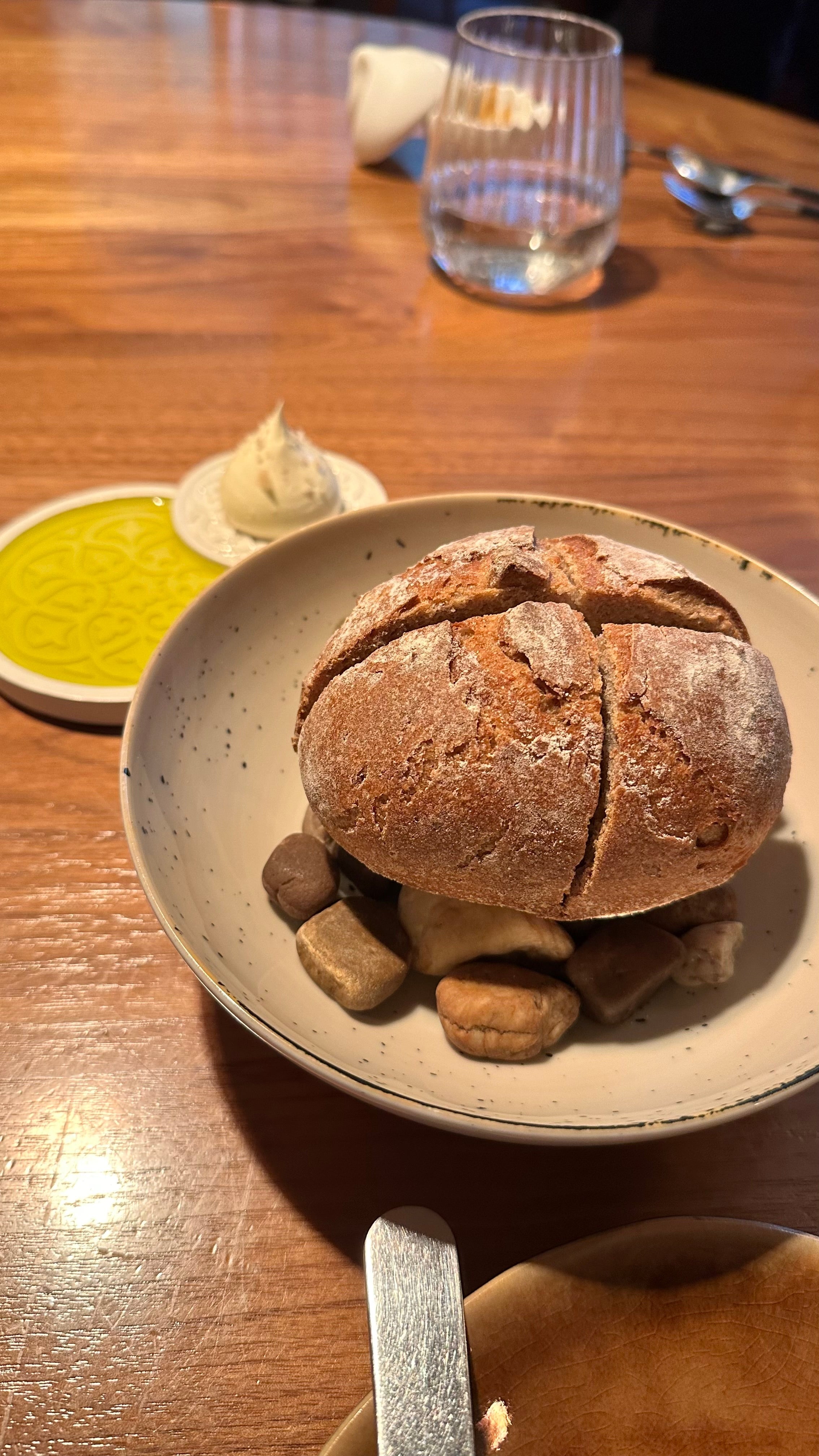 Rustic round loaf of bread served over decorative pebbles in a bowl, with olive oil and butter on the side.
