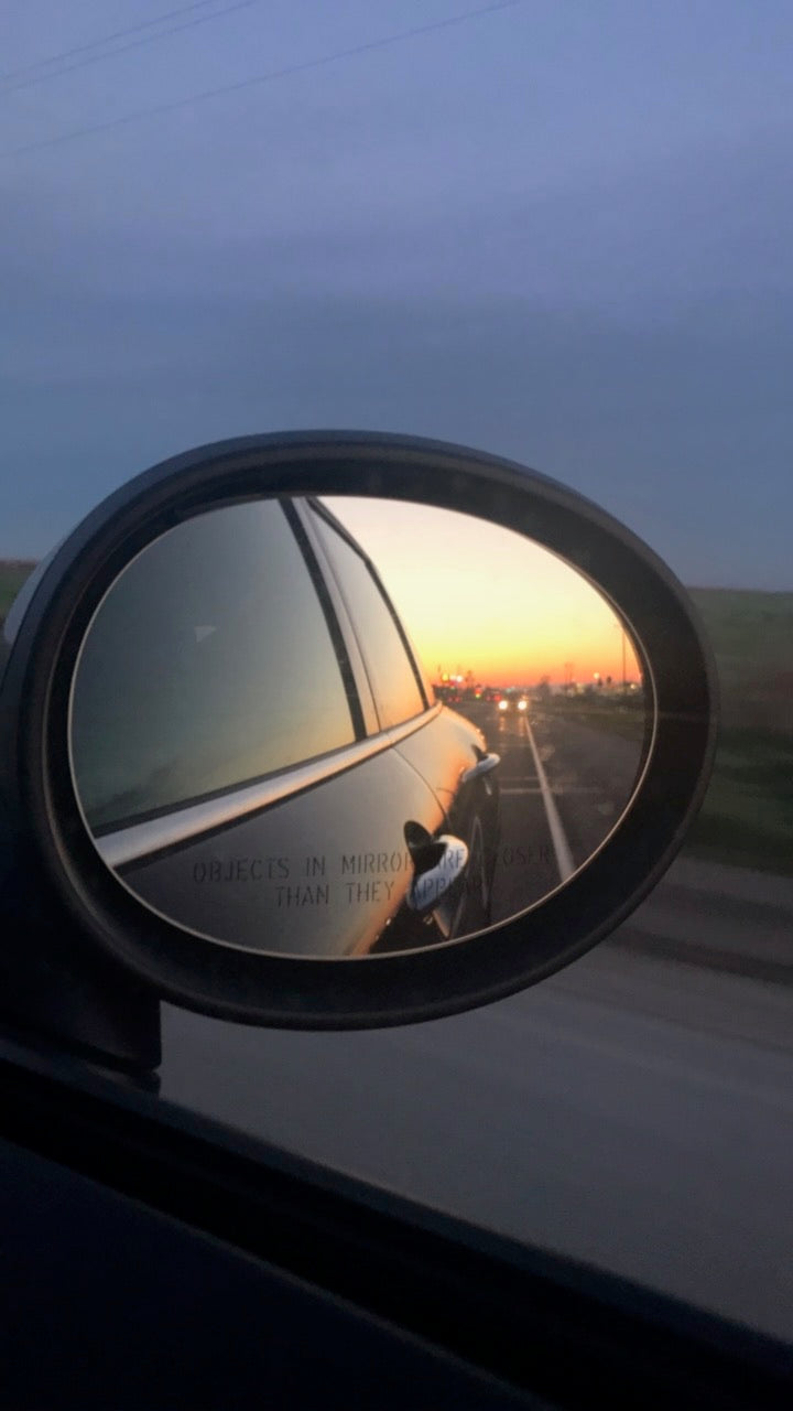 Car side mirror reflecting a sunset view along an open road.