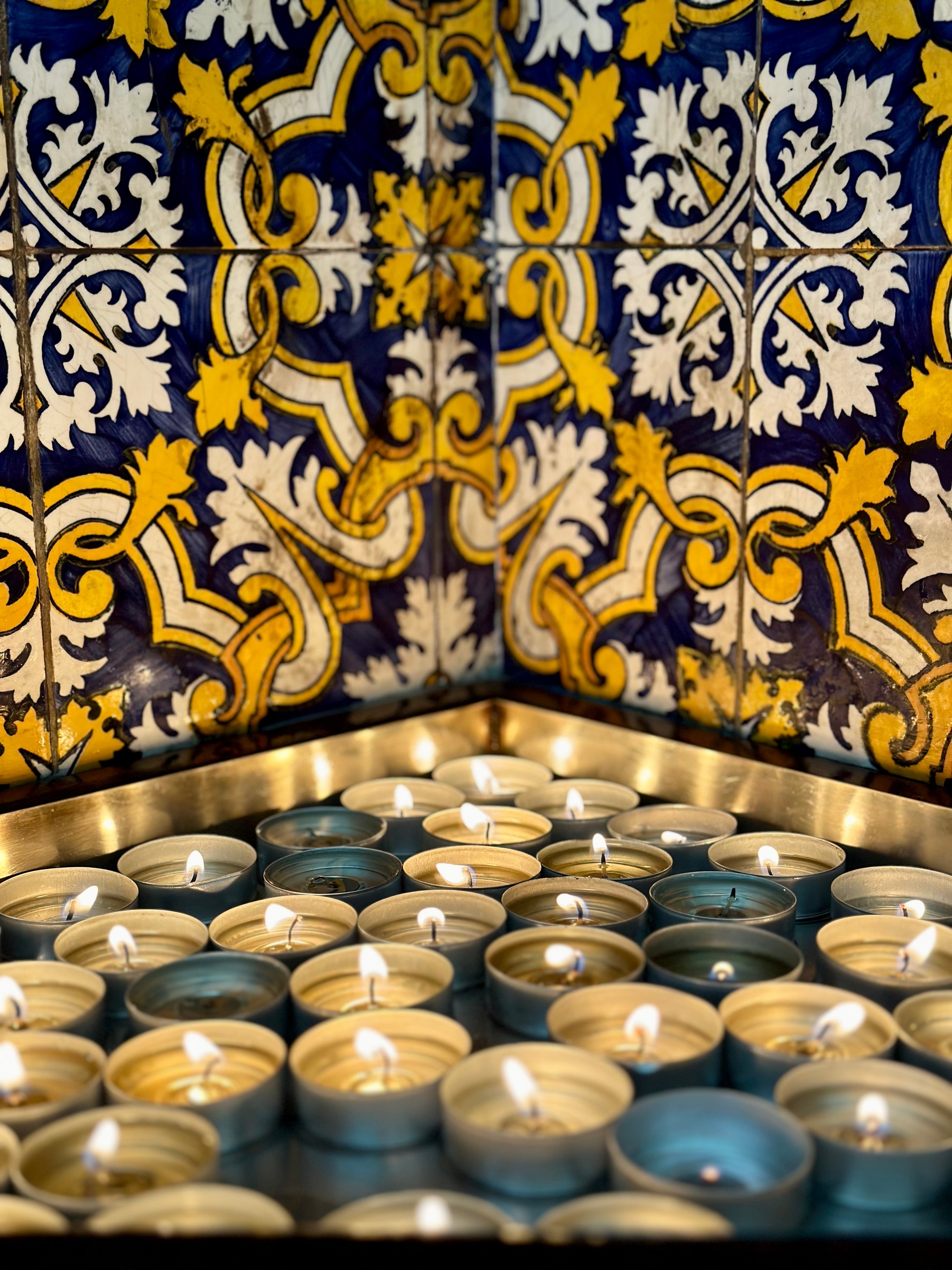 Lit votive candles arranged beneath ornate blue, white, and gold decorative tiles.
