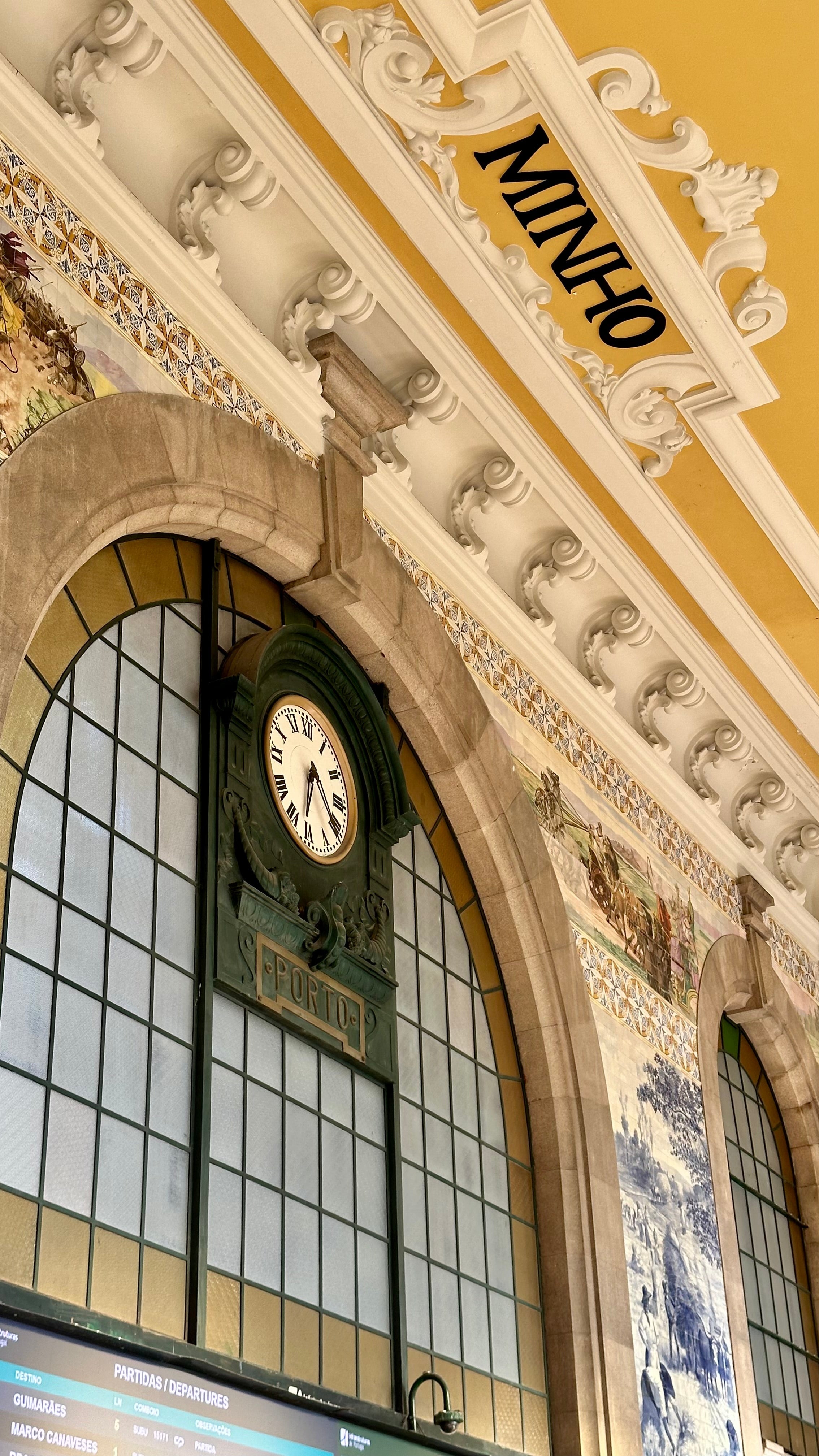 Historic train station facade featuring a large arched window, decorative tilework, and a clock beneath the "MINHO" sign