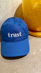 A royal blue baseball cap with the word 'trust' embroidered in white lowercase letters on the front, displayed on a textured beige surface next to a yellow decorative object.