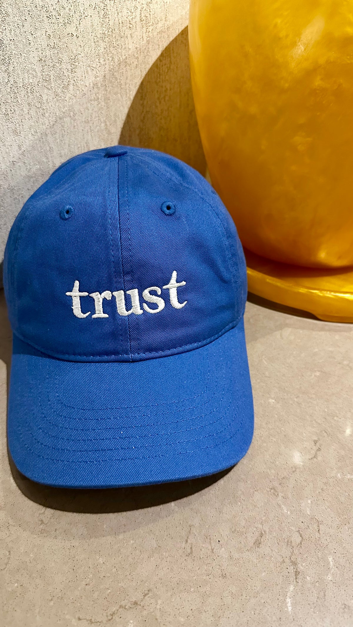 A royal blue baseball cap with the word 'trust' embroidered in white lowercase letters on the front, displayed on a textured beige surface next to a yellow decorative object.