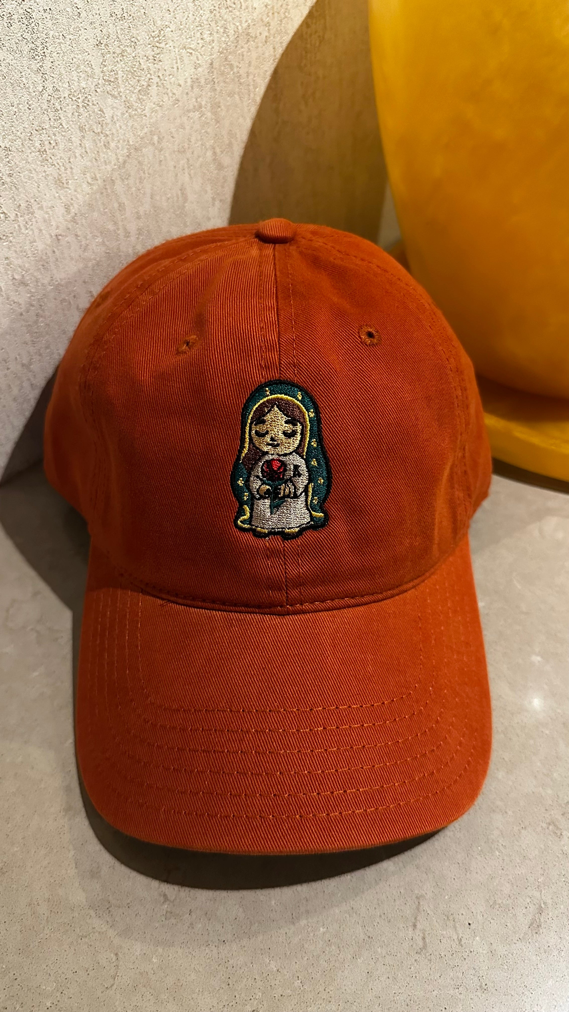 A burnt orange cotton baseball cap featuring an embroidered image of Our Lady of Guadalupe holding a red rose, , resting on a textured beige surface with a yellow decorative object in the background.