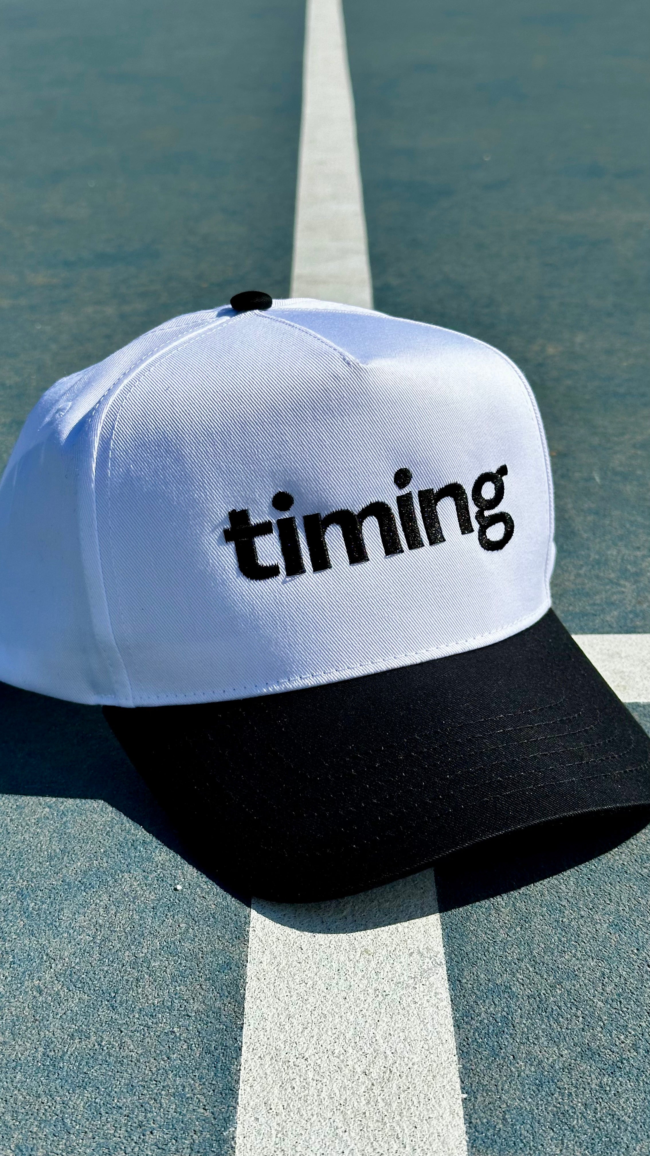 White and black trucker hat with black “timing” embroidery on the front, photographed on an outdoor tennis court.
