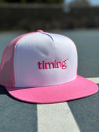Pink and white trucker hat with pink “timing” embroidery and a small yellow sparkle on the front, photographed outdoors.