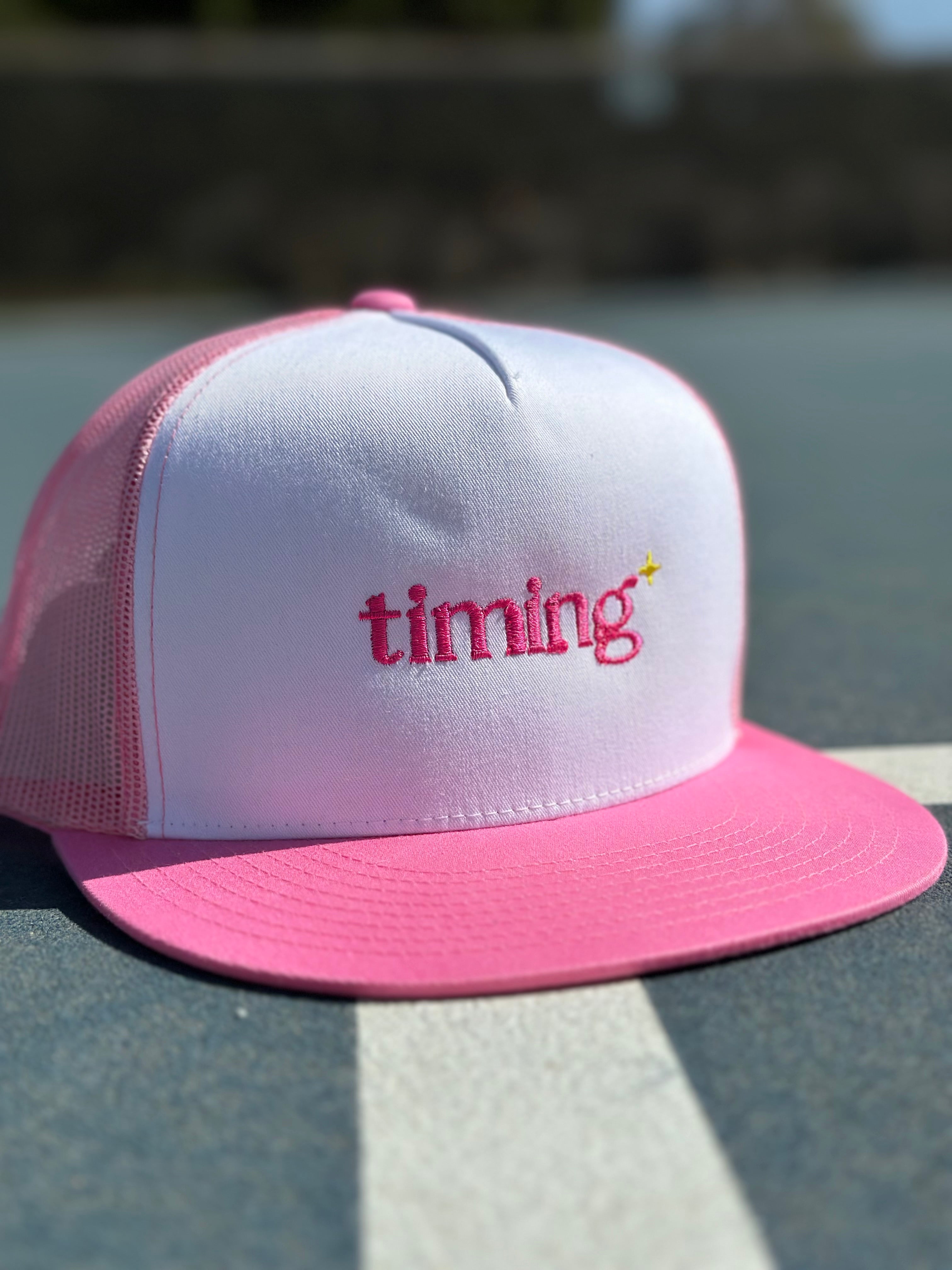 Pink and white trucker hat with pink “timing” embroidery and a small yellow sparkle on the front, photographed outdoors.