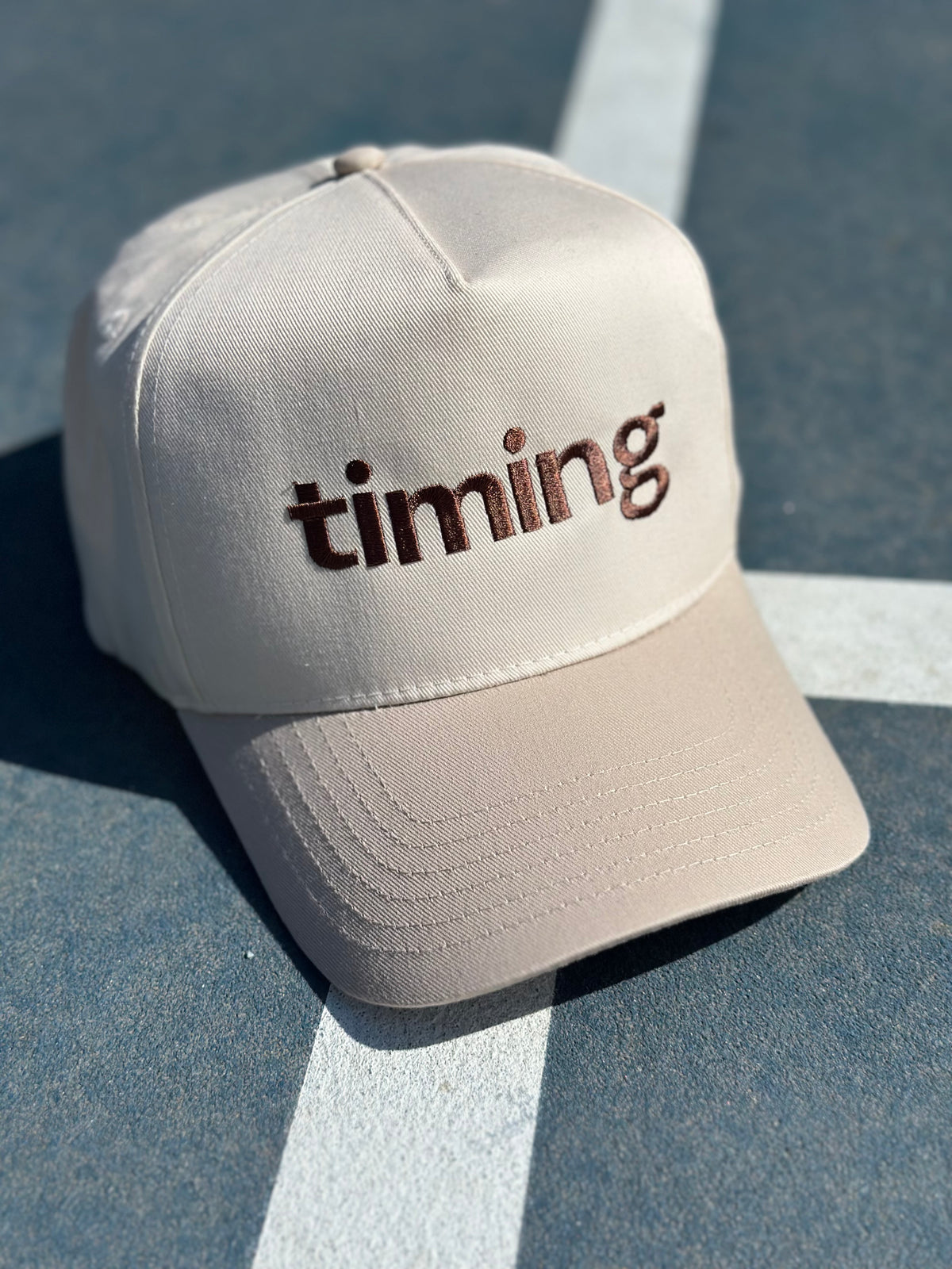 Cream baseball cap with dark brown “timing” embroidery on the front, photographed on an outdoor tennis court.