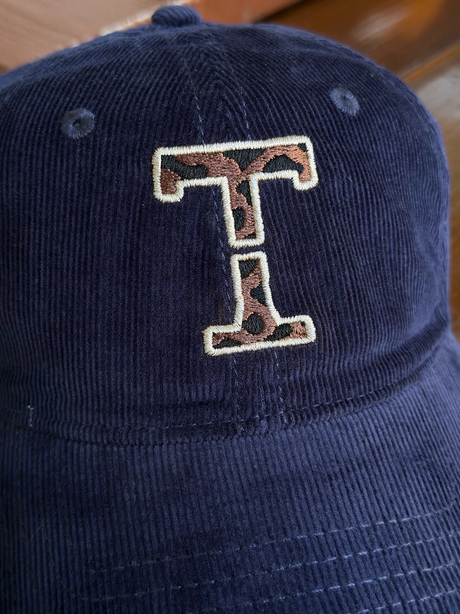 Close-up of the leopard-print Threshold T embroidery with a cream outline and intentional center gap on a navy corduroy cap.