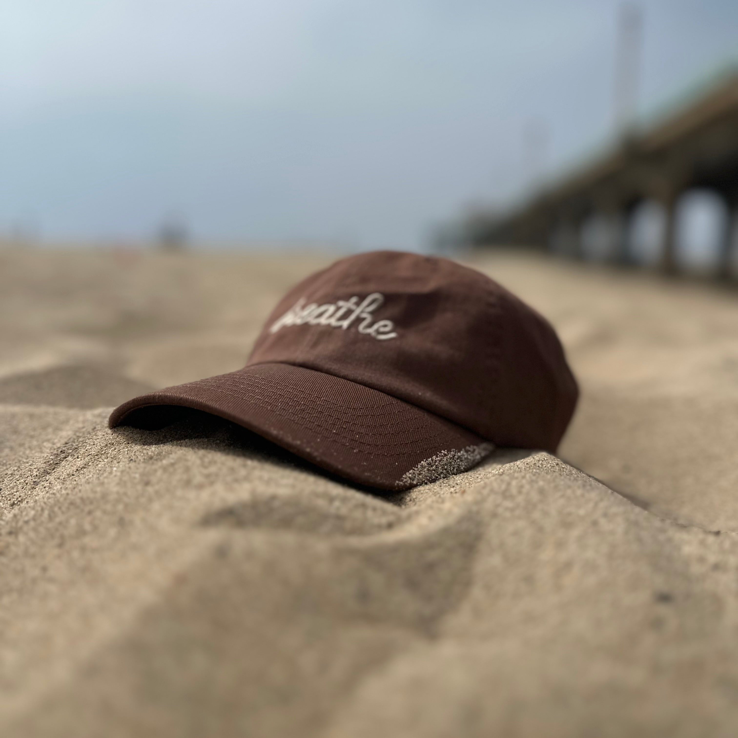 Brown dad hat featuring embroidered "breathe" script resting on the sand at the beach.