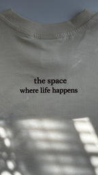 Back view of a cream cotton T-shirt featuring the phrase "the space where life happens" printed in burgundy lettering.