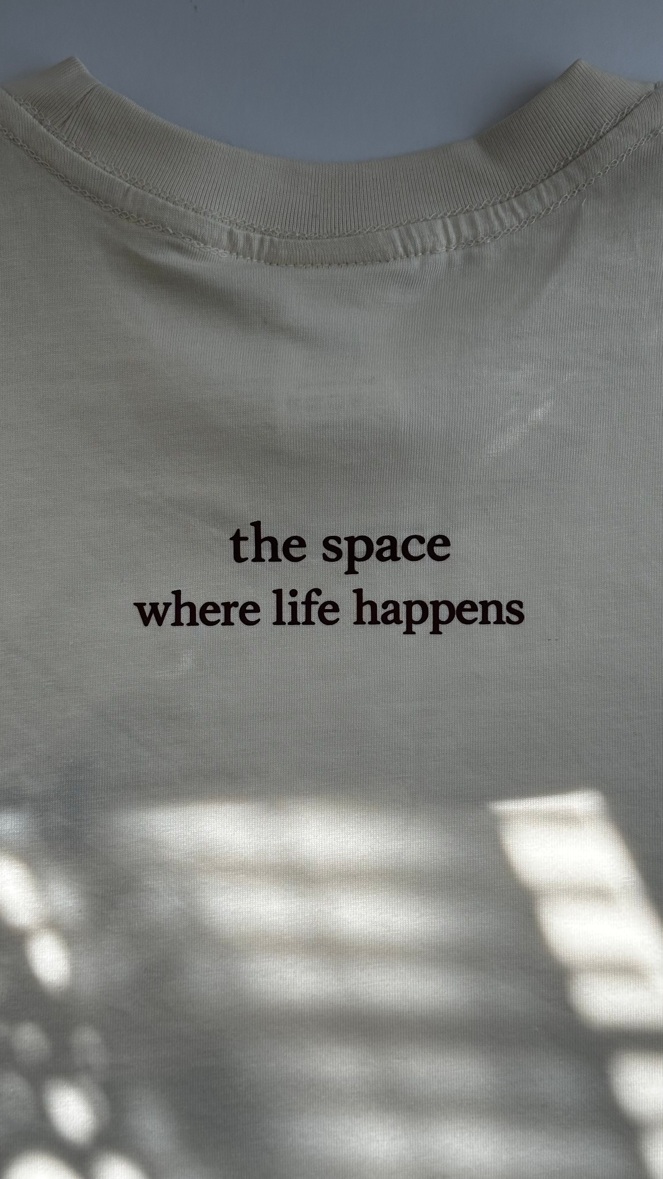 Back view of a cream cotton T-shirt featuring the phrase "the space where life happens" printed in burgundy lettering.