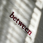 Close-up of a cream cotton T-shirt featuring "between" embroidered in burgundy script, photographed in natural light.