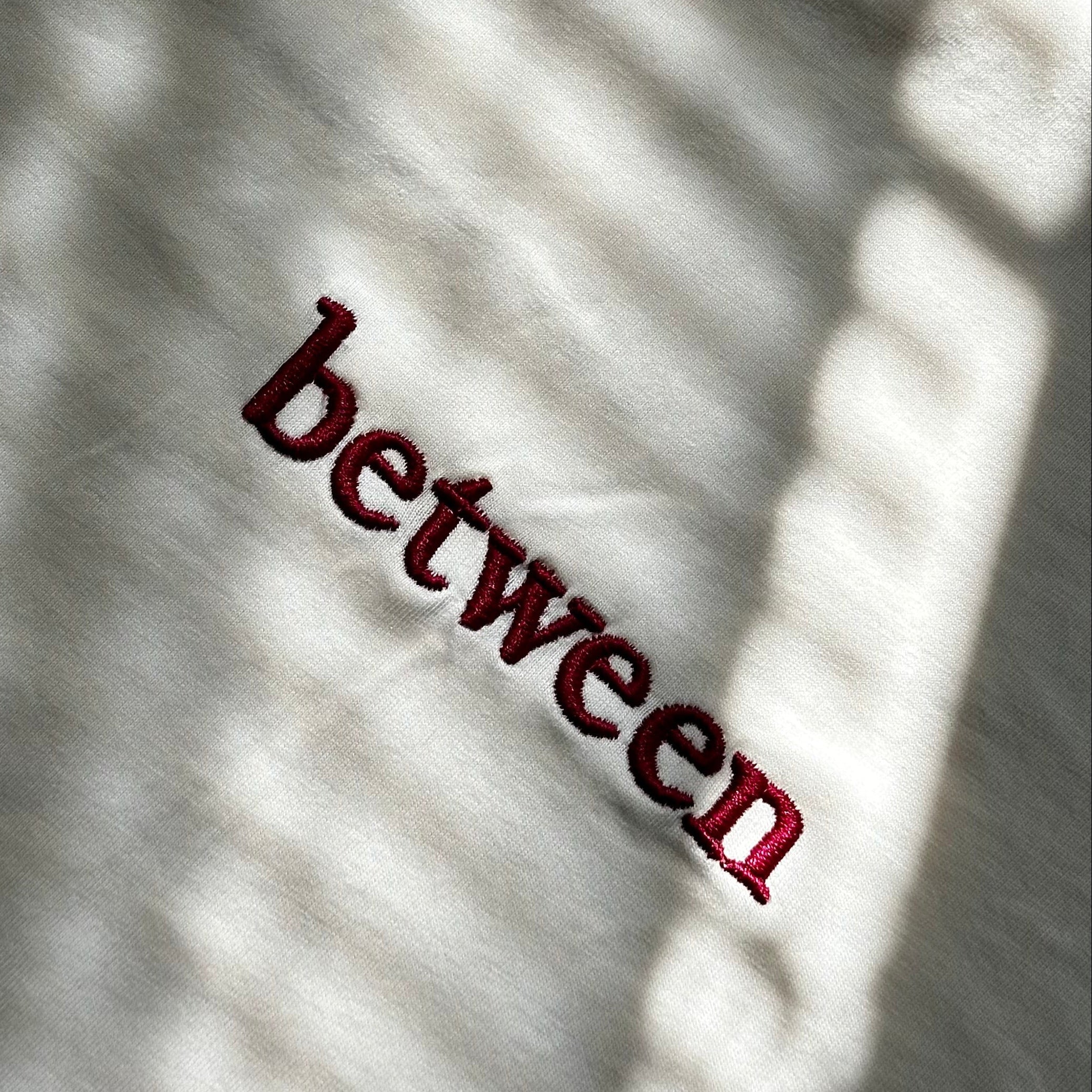 Close-up of a cream cotton T-shirt featuring "between" embroidered in burgundy script, photographed in natural light.