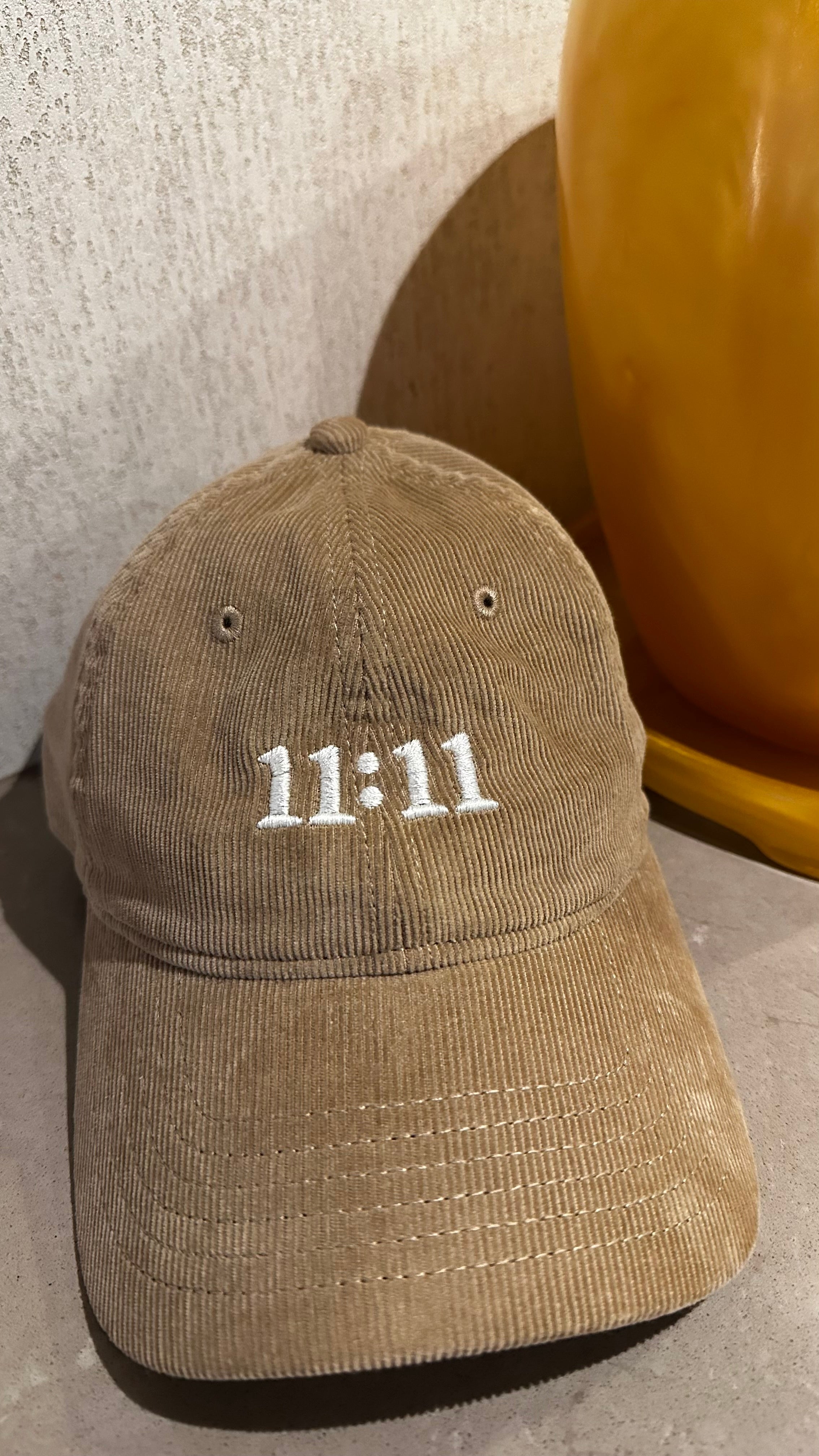 A beige corduroy baseball cap with '11:11' embroidered in white on the front, resting on a tan surface next to a yellow decorative object.