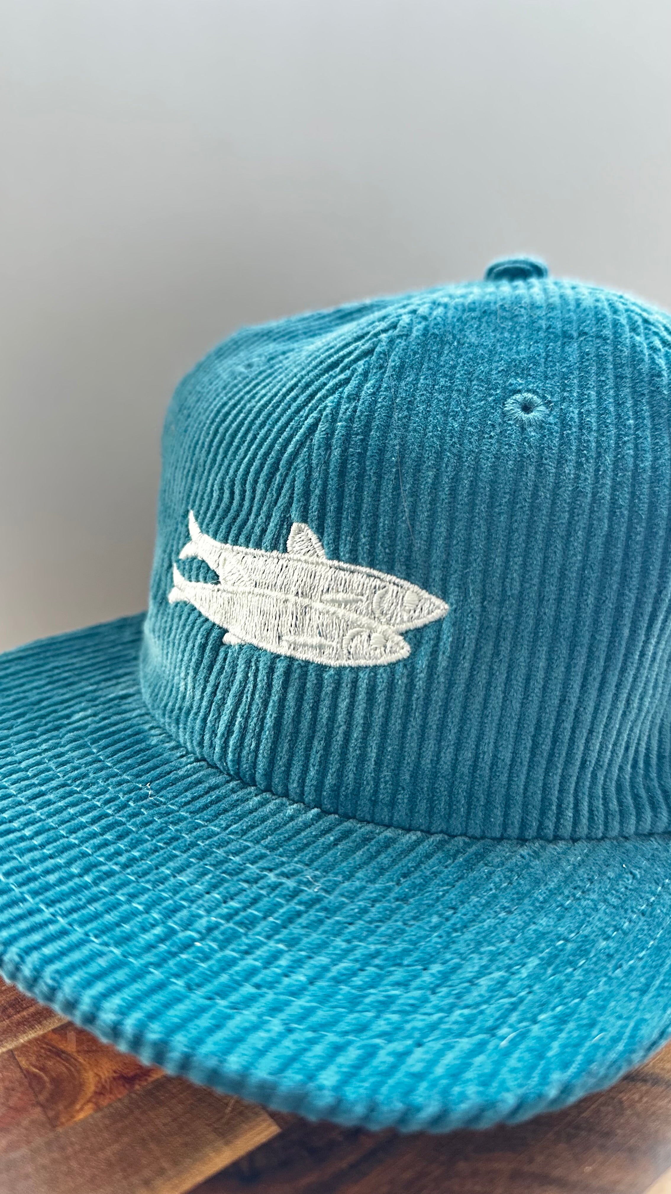 Angled view of a teal corduroy hat featuring ivory sardine embroidery