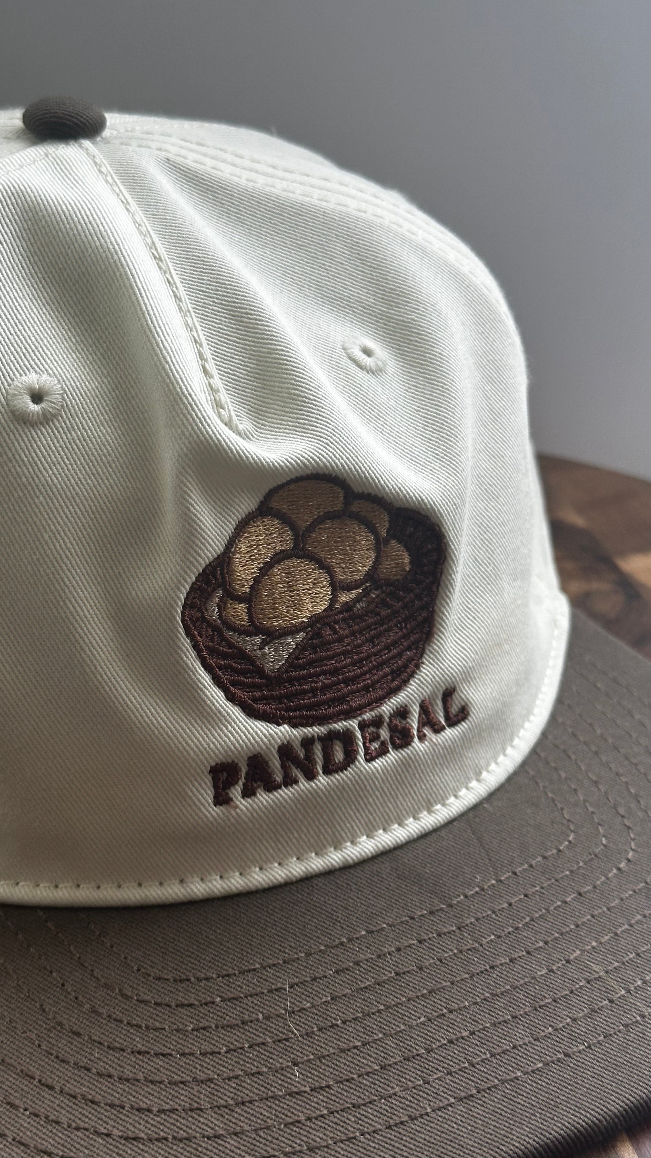 Angled close-up of a cream and brown two-tone cap with embroidered basket of pandesal and "PANDESAL" text.