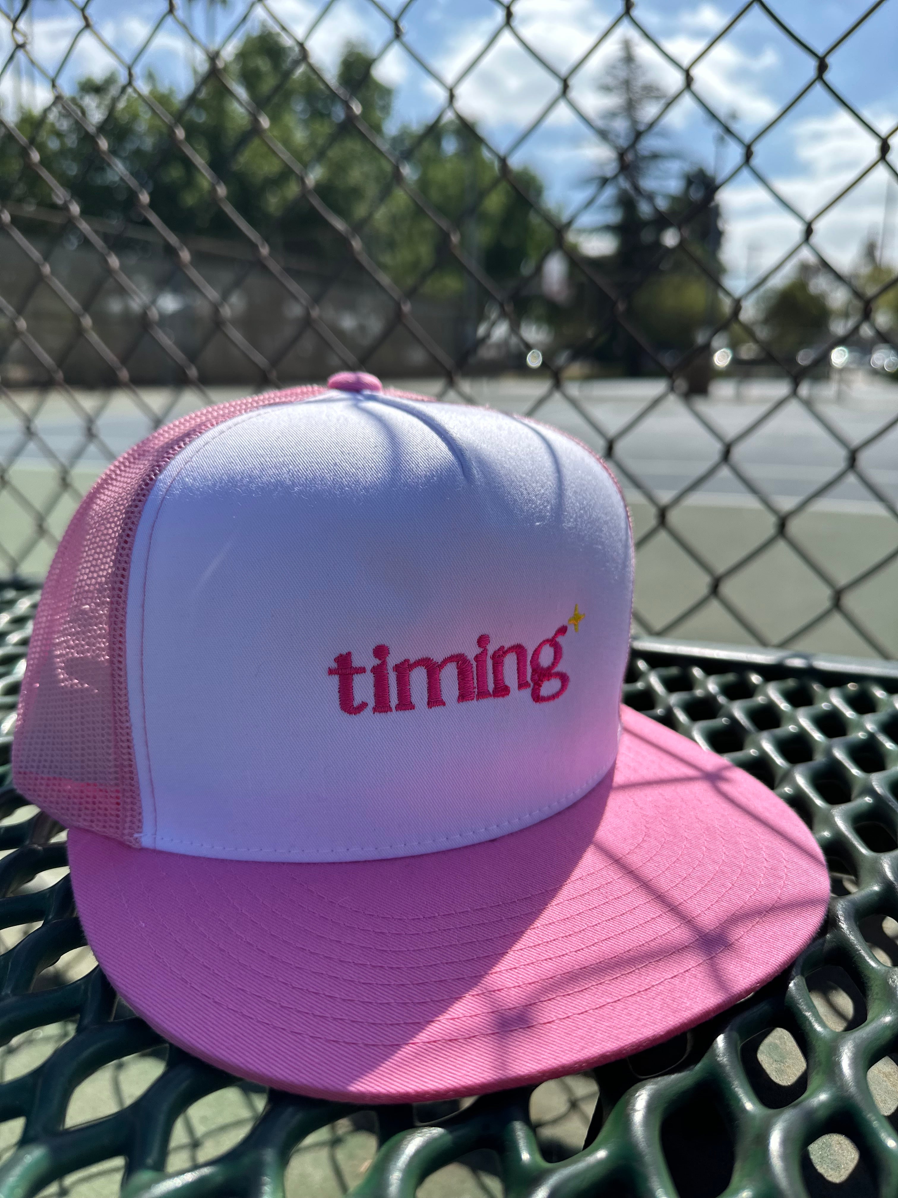 Pink and white trucker hat with pink “timing” embroidery and a small yellow sparkle, photographed courtside at an outdoor tennis court.