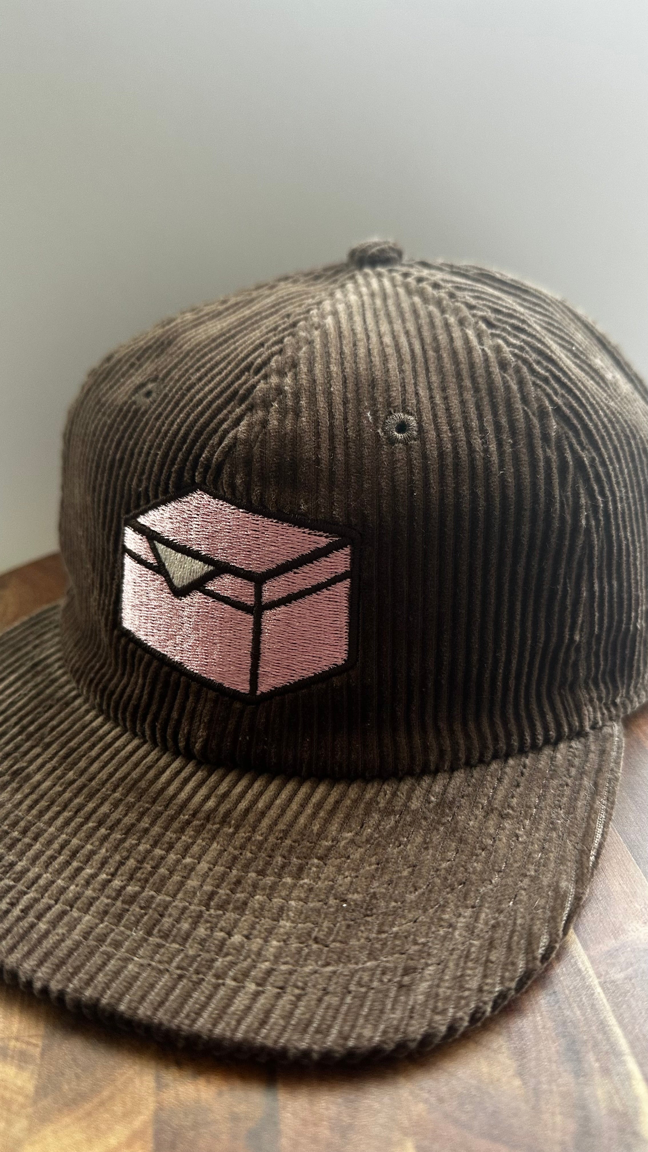 Brown corduroy hat embroidered with a pink donut box icon on a wooden surface.