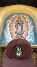 Rose Protector embroidered hat displayed beneath a mural of Our Lady of Guadalupe at a church entrance.