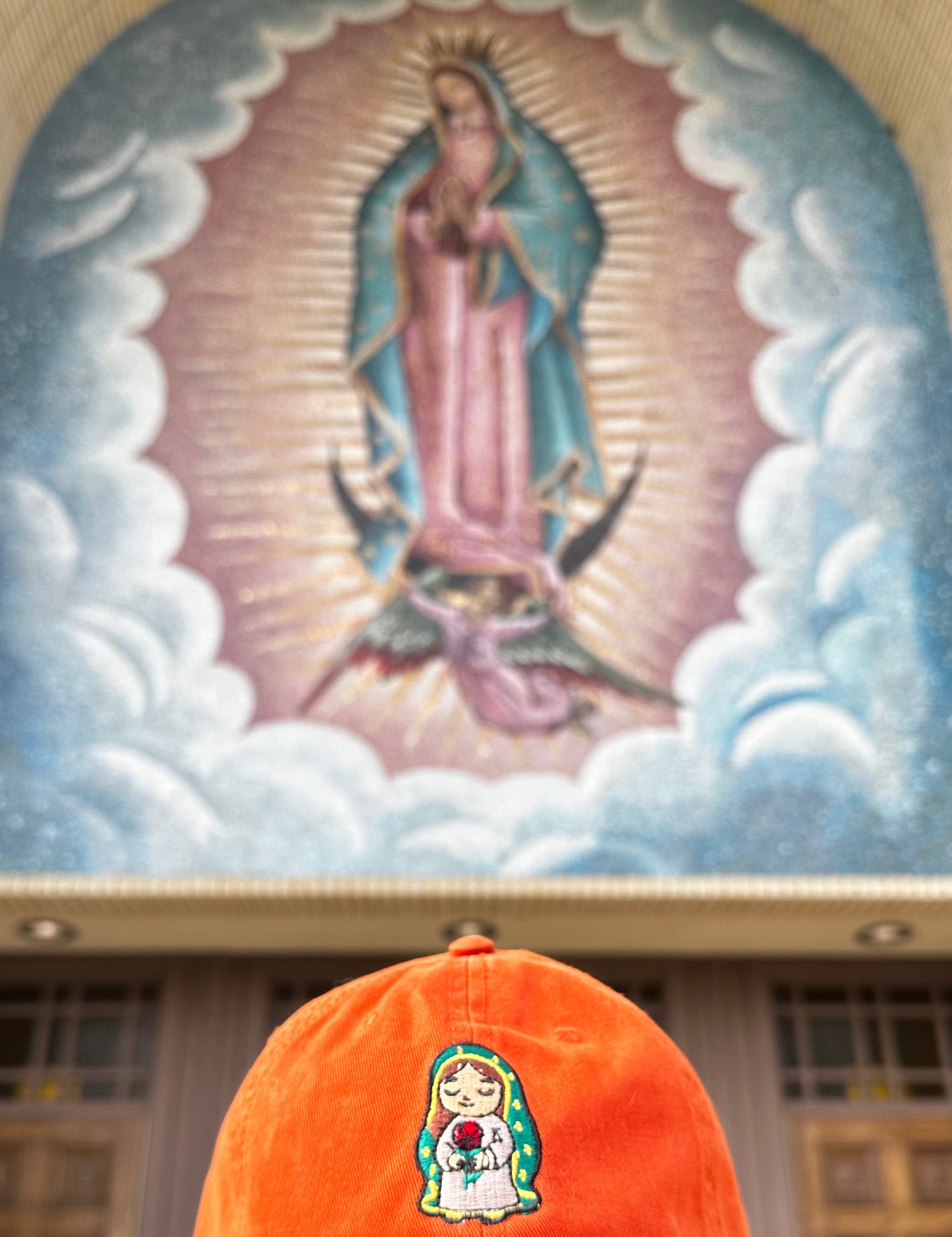 Person wearing a burnt orange corduroy baseball cap featuring an embroidered image of Our Lady of Guadalupe holding a red rose, photographed in front of a softly blurred mural of Our Lady of Guadalupe.