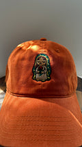 Front view of a burnt orange corduroy baseball cap featuring an embroidered image of Our Lady of Guadalupe holding a red rose, photographed in natural sunlight.