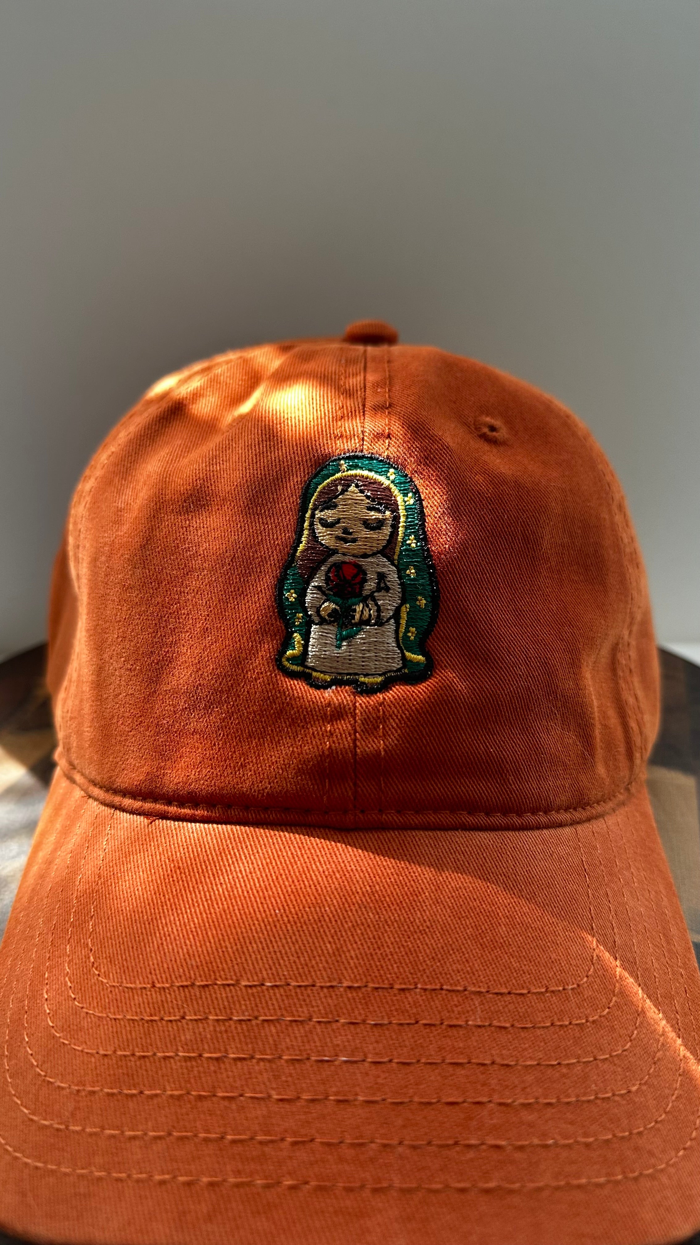 Front view of a burnt orange corduroy baseball cap featuring an embroidered image of Our Lady of Guadalupe holding a red rose, photographed in natural sunlight.