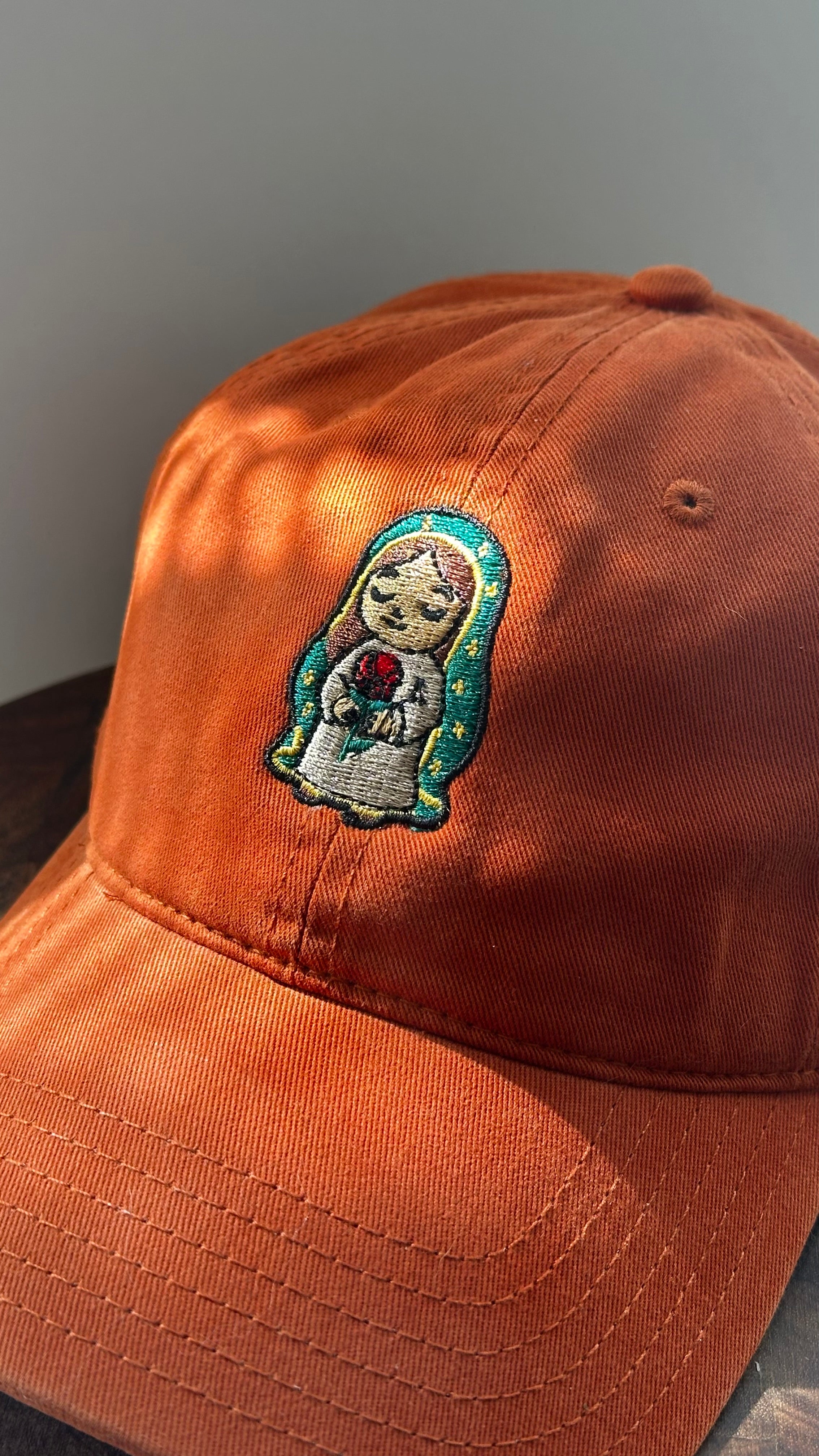 Angled close-up of a burnt orange corduroy baseball cap featuring an embroidered image of Our Lady of Guadalupe holding a red rose, photographed in natural sunlight.