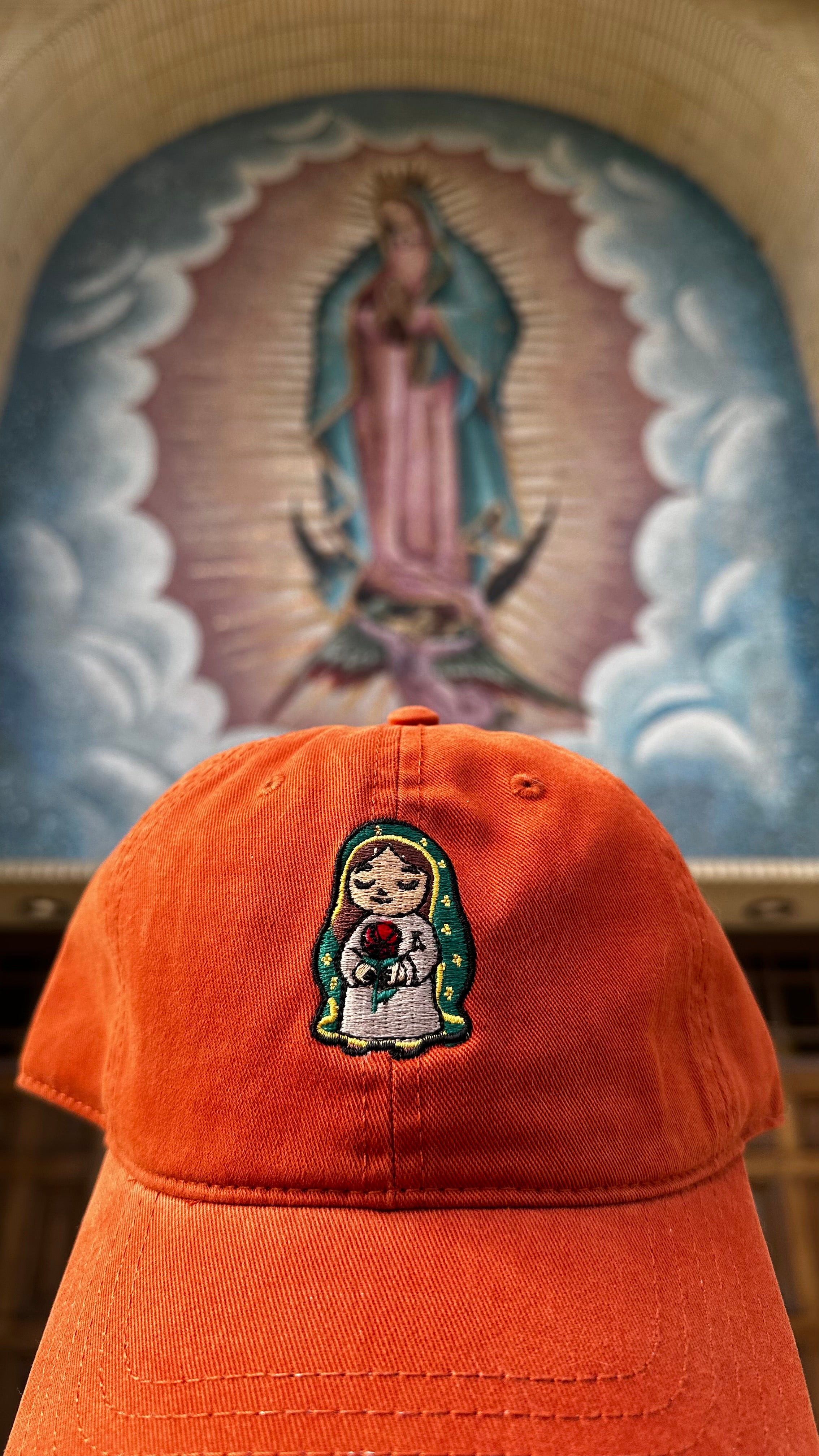 Front view of the Rose Protector embroidered Burnt Orange dad cap, photographed inside a church with a softly blurred image of Our Lady of Guadalupe in the background.