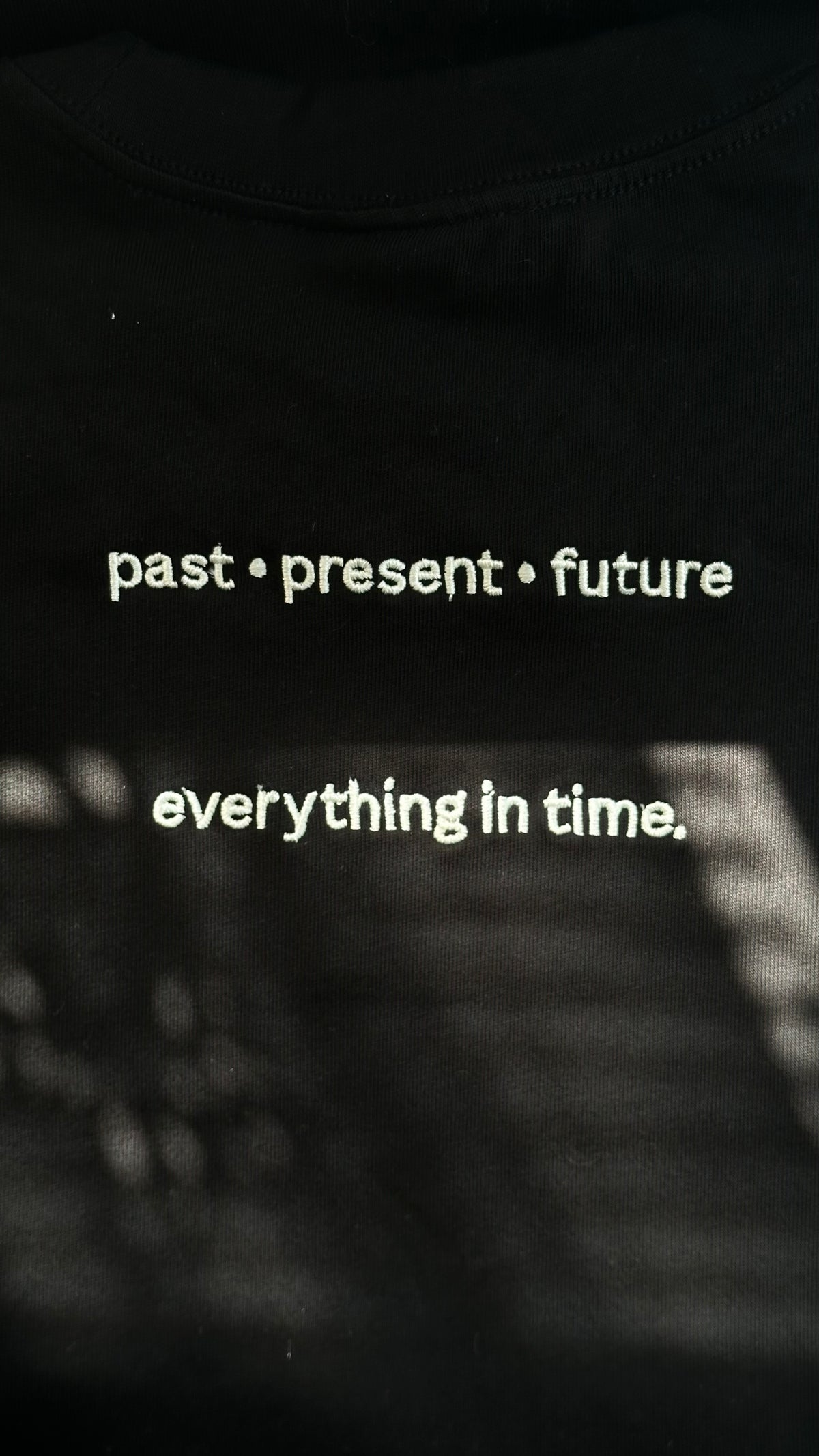 Back view of a black cotton T-shirt featuring the phrases "past • present • future" and "everything in time." embroidered in ivory lettering.