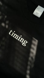 Close-up of a black cotton T-shirt featuring "timing" embroidered in ivory lettering at the center chest, photographed in natural light.
