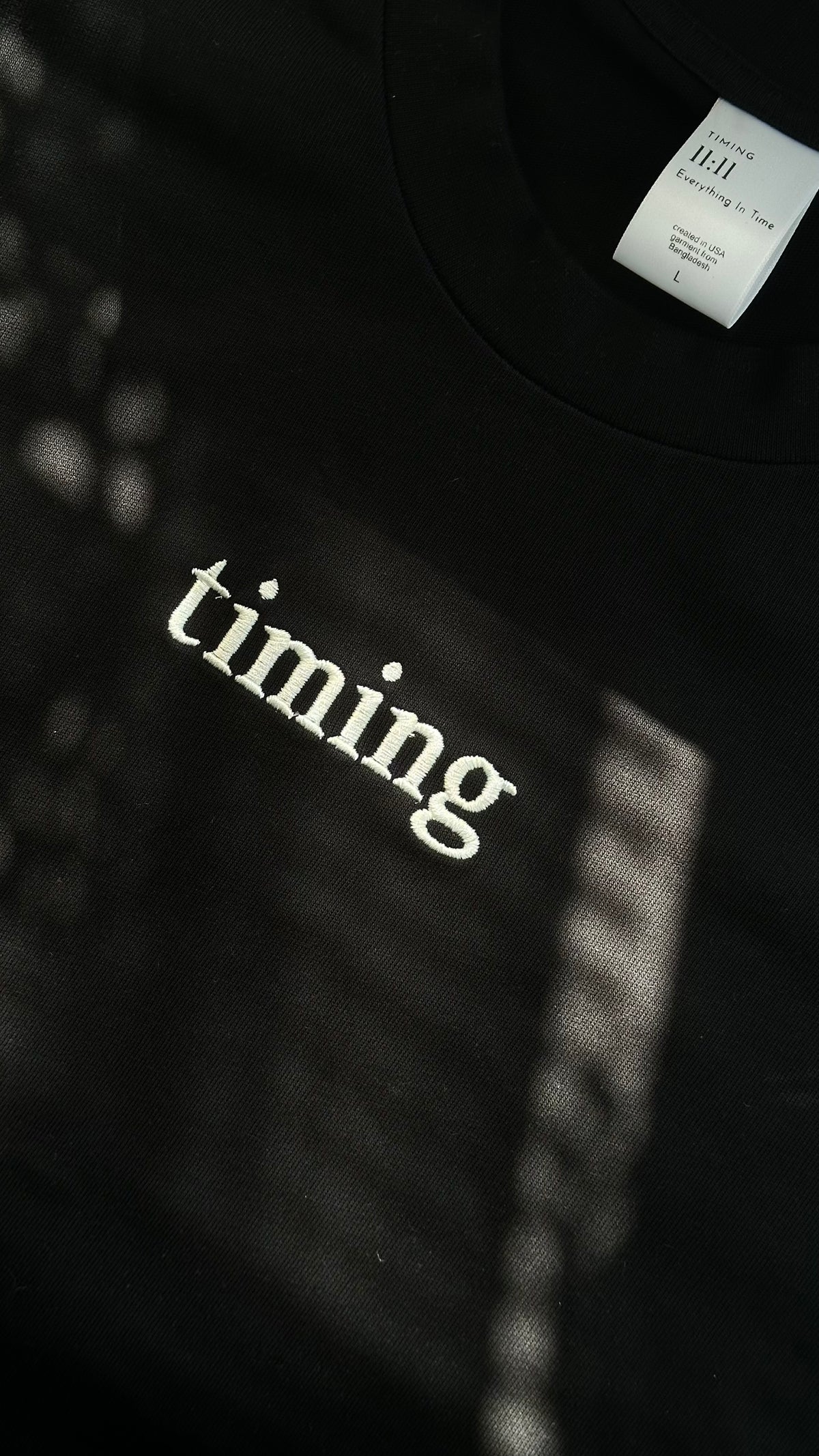 Close-up of a black cotton T-shirt featuring "timing" embroidered in ivory lettering at the center chest, photographed in natural light.