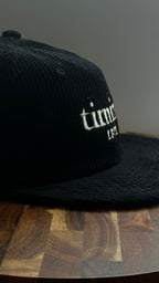 Angled view of a black corduroy hat featuring the embroidered "timing 11:11" logo in ivory.