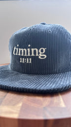 Angled view of a blue corduroy hat featuring the embroidered "timing 11:11" logo in ivory.