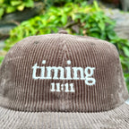 Close-up of a brown corduroy hat featuring the embroidered "timing 11:11" logo in ivory