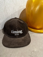 Brown corduroy hat featuring ivory "timing 11:11" embroidery, styled beside an amber vase on a stone tabletop.