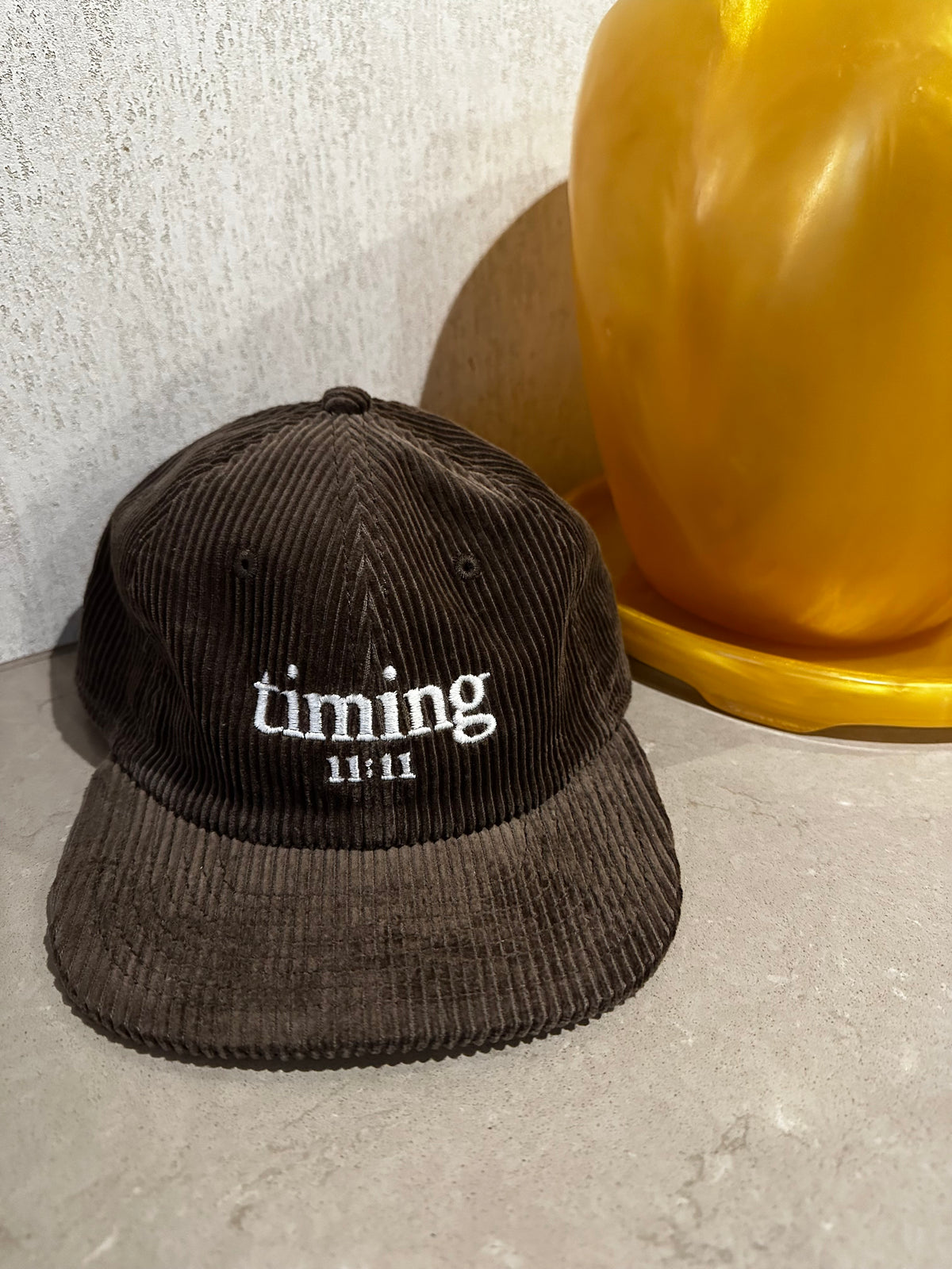 Brown corduroy hat featuring ivory "timing 11:11" embroidery, styled beside an amber vase on a stone tabletop.