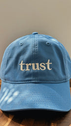 Light blue cotton dad hat embroidered with the word "trust." in cream thread, photographed from the front on a wooden surface.