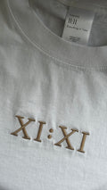 Close-up of the embroidered XI:XI graphic and Timing 11:11 signature label on a white t-shirt.