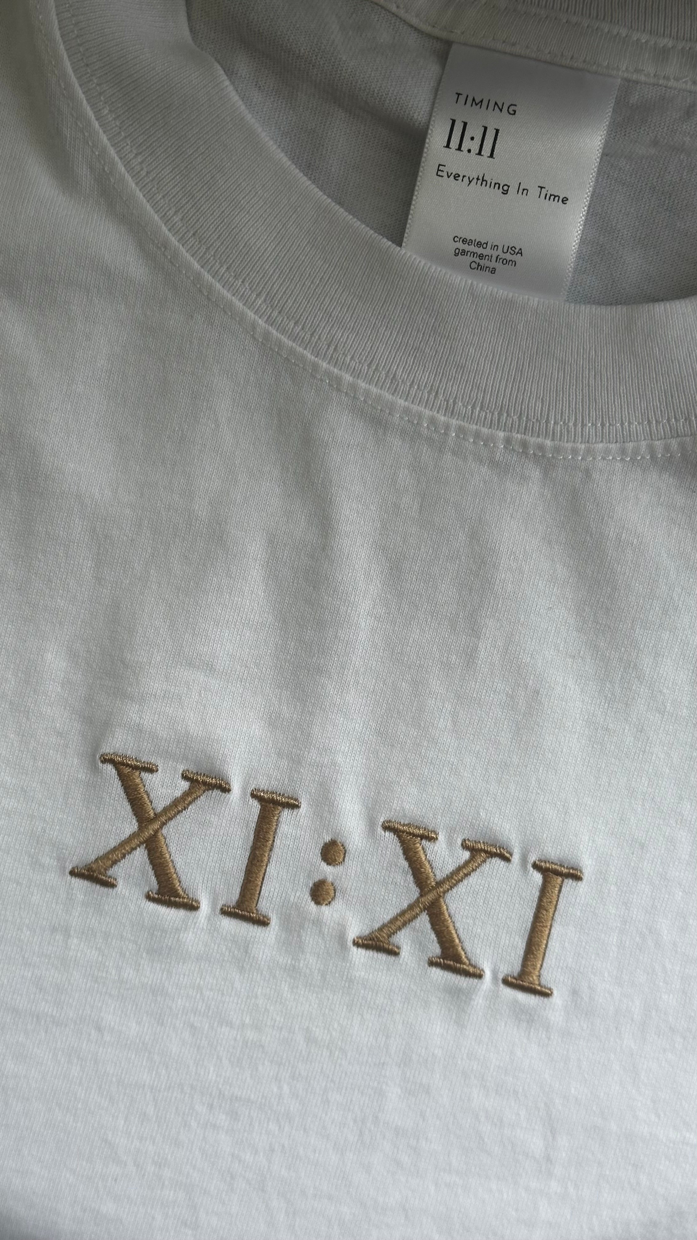 Close-up of the embroidered XI:XI graphic and Timing 11:11 signature label on a white t-shirt.